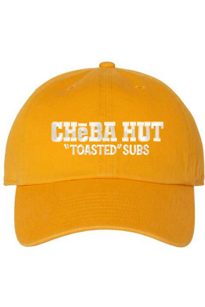 A gold baseball cap with a curved brim. The front of the cap features the white stylized text 'CHEBA HUT "TOASTED" SUBS' embroidered across the center. The cap has a visible button on the top and an adjustable strap in the back (not visible).