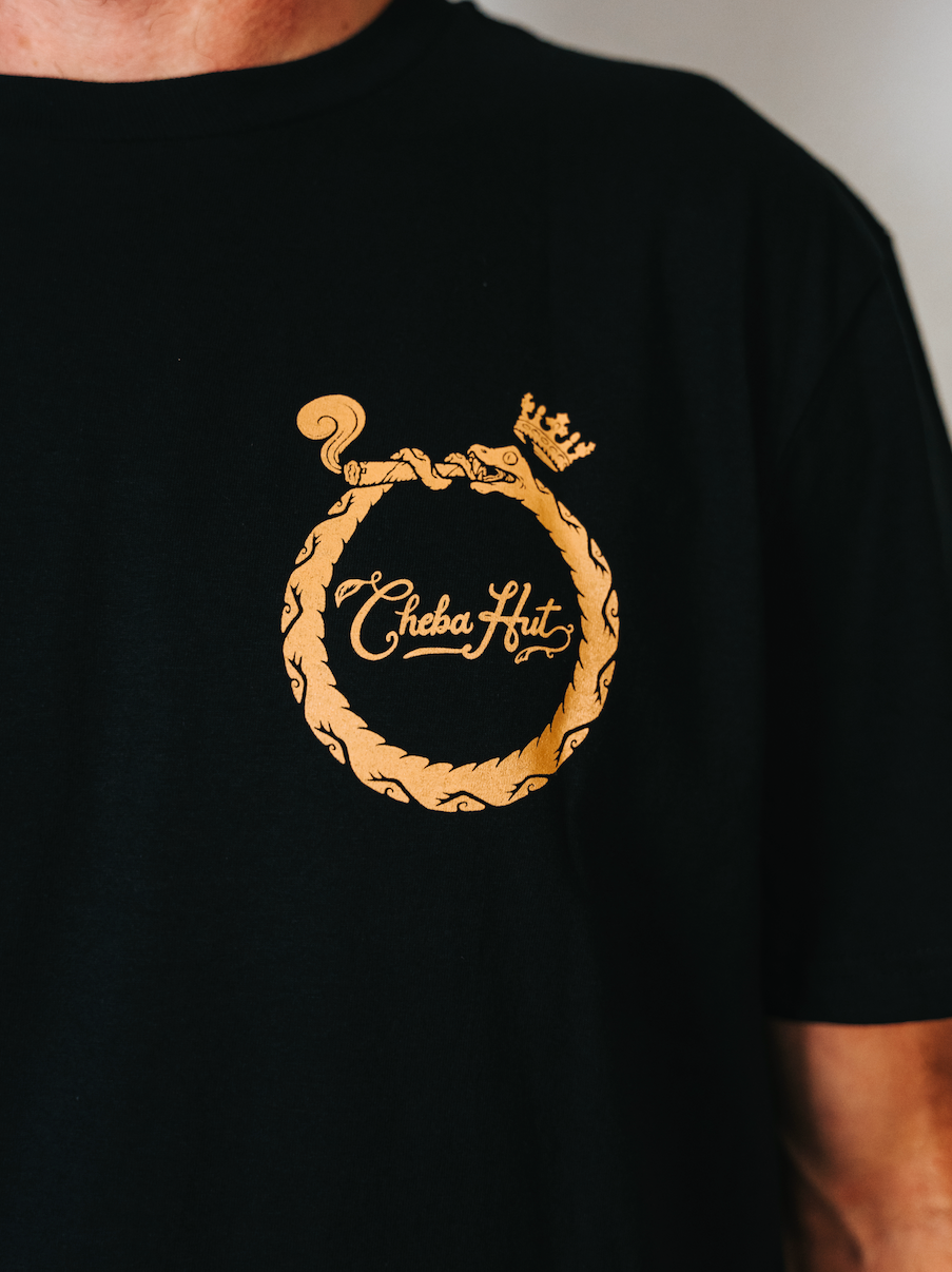 Front image of the Cheba Hut Secret tee features a black tee shirt with an image of a snake, smoking a joint, circled around the words Cheba Hut on the left side of the shirt. 